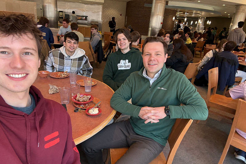Students take a selfie and smile with Trevor Peters while eating lunch in a dining court. 