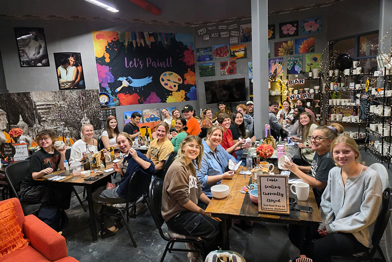 A large group of students smile as they paint ceramic creations in a colorful setting. 